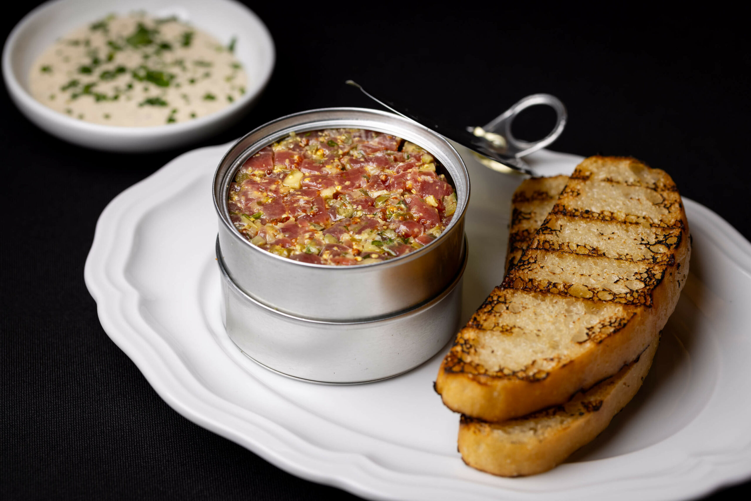 A close-up side view of the tartare tin, paired with grilled bread and a creamy herb dip.