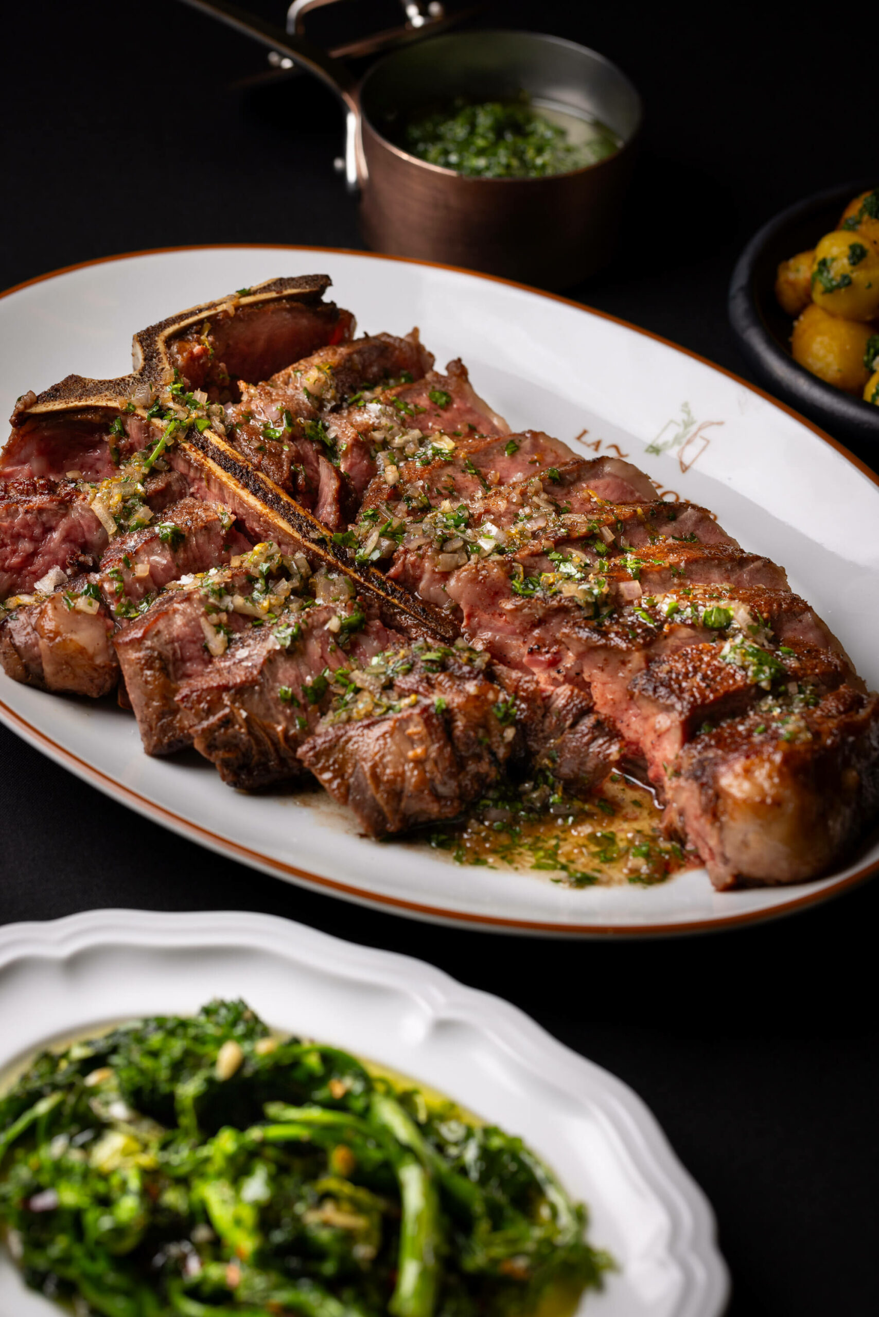 A grilled porterhouse steak served with herb gremolata, accompanied by sides of green vegetables and roasted potatoes.