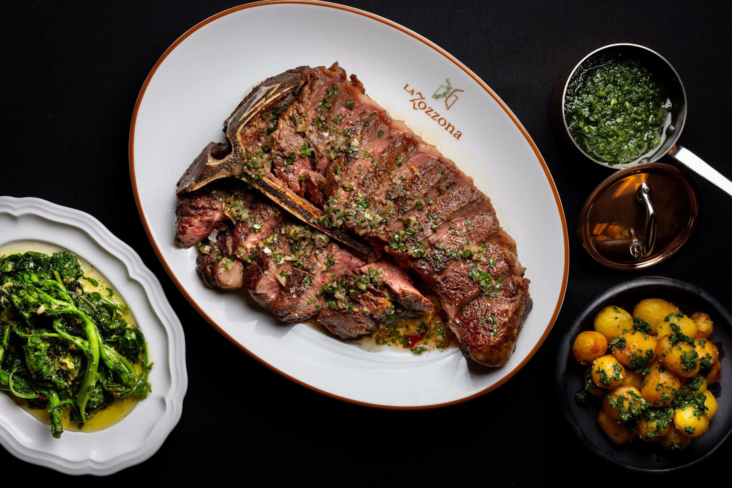 An overhead shot of the porterhouse steak paired with roasted potatoes and green vegetables, along with a small bowl of herb sauce.