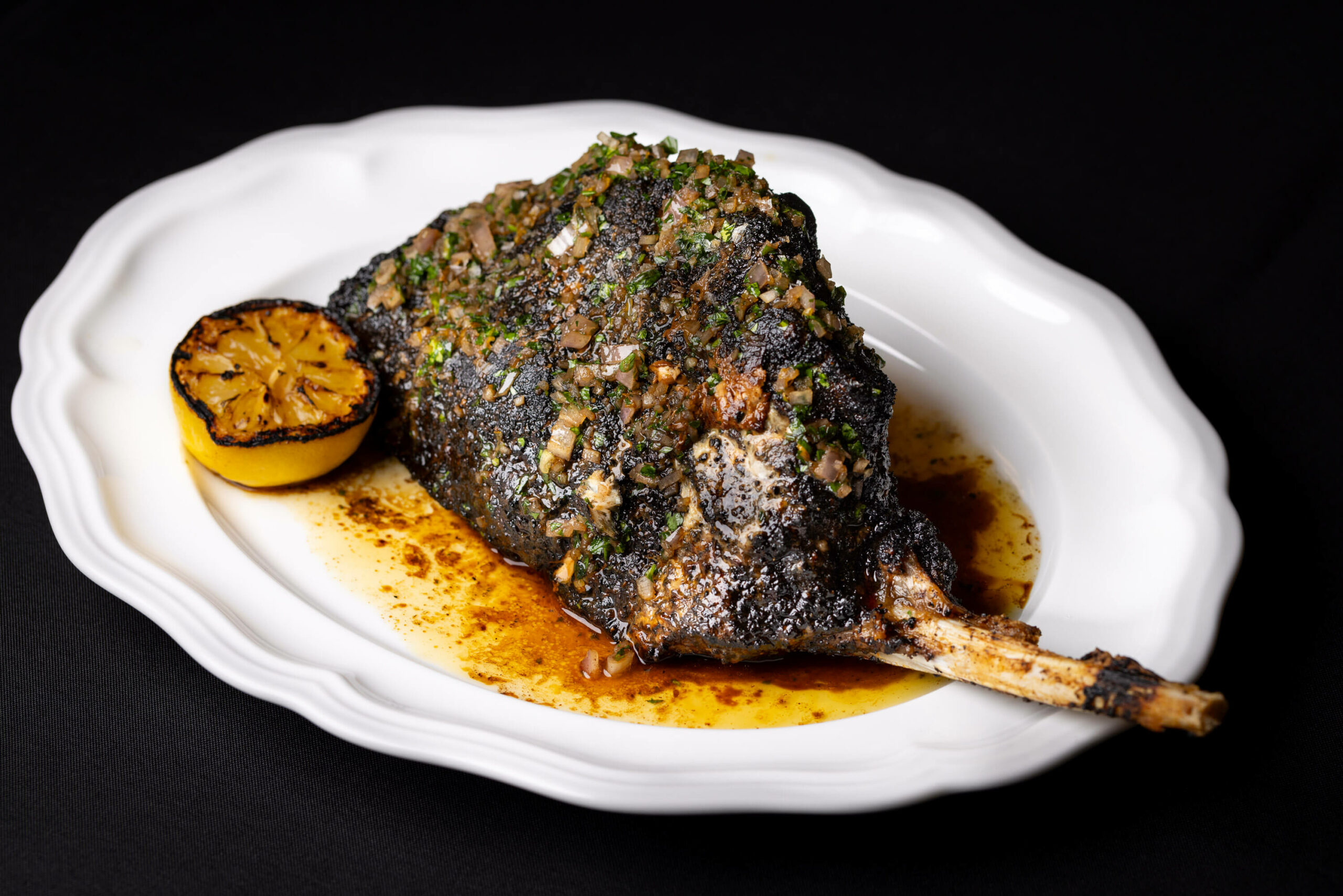 A lamb chop served with a charred lemon half, topped with fresh herbs and olive oil on a white plate.