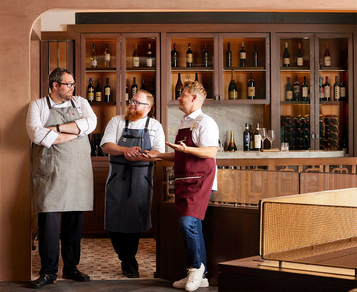 Three chefs, including Richard Blais, sharing a light moment in front of the wine display.