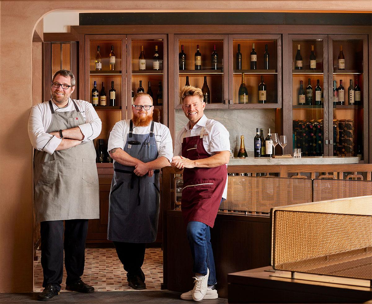 Chef Richard Blais with two fellow chefs, smiling inside the warmly lit wine bar area.